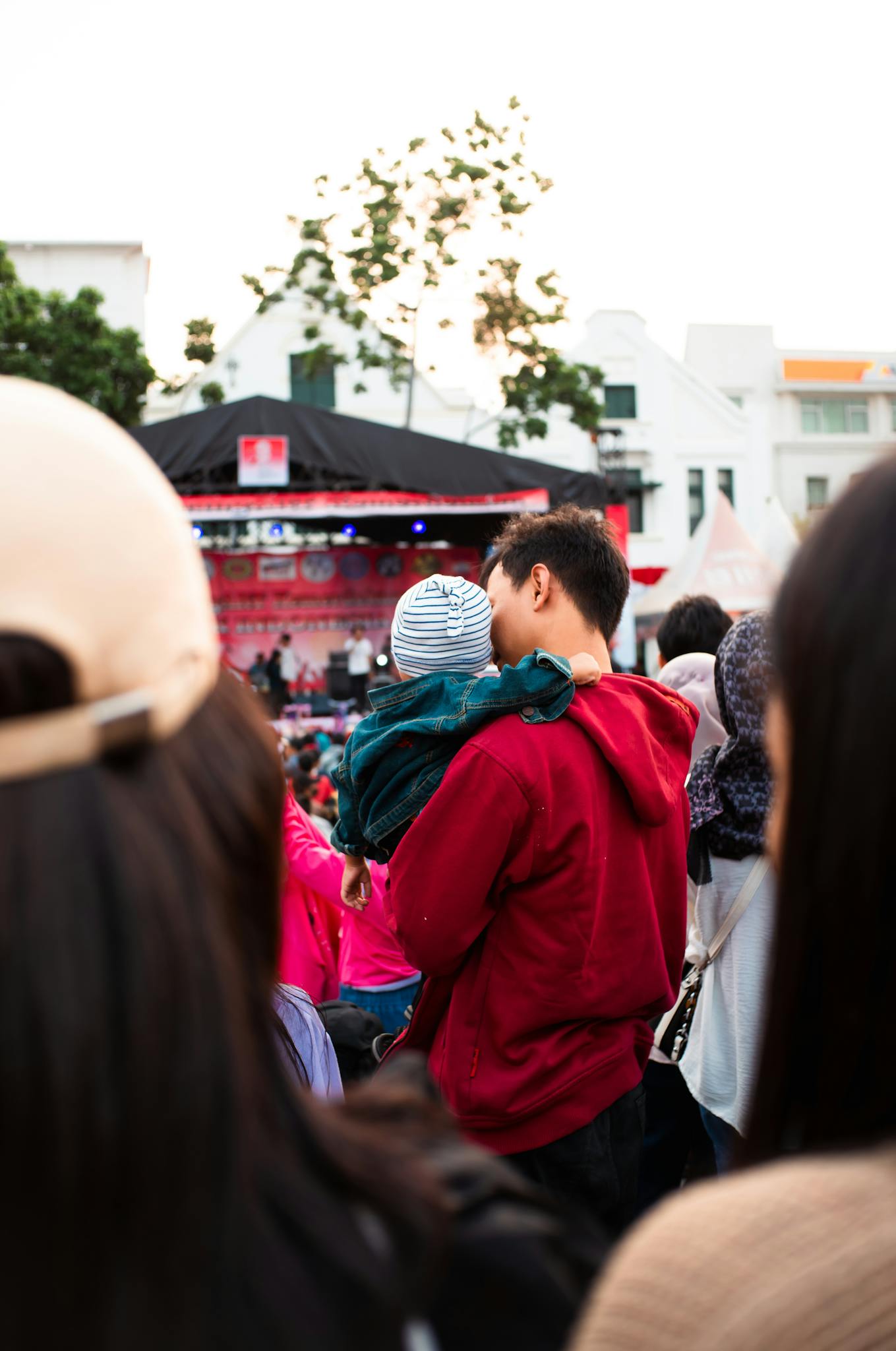 Father in red jacket holding child at an outdoor concert surrounded by a crowd.